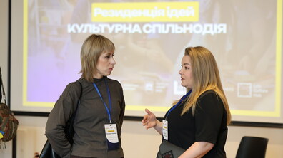 Дві учасниці резиденції спілкуються під час події. Вони стоять у залі перед великим екраном, на якому видно напис «Резиденція ідей. Культурна спільнодія». Обидві учасниці мають бейджі, а одна з них жестикулює під час розмови.