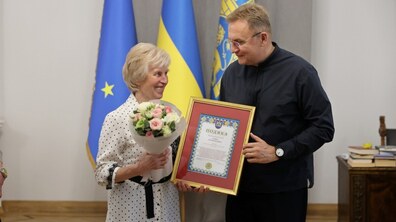 Мер Львова Андрій Садовий нагороджує подякою від громади Львова переможницю конкурсу «Найкращий двірник Львова-2026» Ганну Петричкович. Жінка усміхається, тримаючи букет квітів у світлих тонах.