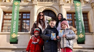 П'ятеро людей стоять біля Різдвяної пошти: двоє усміхнених жінок та троє дітей, позаду фасад палацу Бандінеллі, площа Ринок 2, прикрашений святковими банерами.