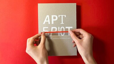 На яскраво-червоному фоні лежить сіра книга з великим білим написом «АРТ БРЮТ» та підзаголовком «мистецтво нейрорізноманітних». Дві жіночі руки обережно тримають тонку червону стрічку-закладку, що горизонтально перетинає обкладинку.