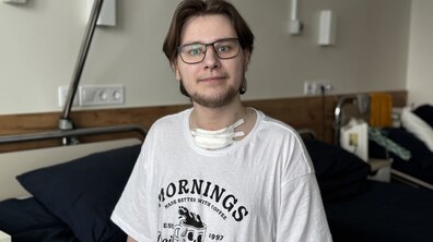 Пацієнт Іван сидить на лікарняному ліжку, на шиї у нього – бинт, заліплений пластиром; хлопець одягнений у білу футболку із принтом чорного кольору
