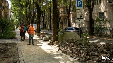 Працівники у спецформі стоять на частково вкритій бруківкою пішохідній алеї. Обабіч бруківка, яку лише планують укладати. Вздовж алеї ростуть високі зелені дерева