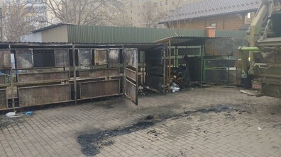 Контейнерний майданчик для збору відходів після пожежі. Уздовж майданчика стоїть металева конструкція з відсіками для сміття — її стінки й дверцята почорнілі від кіптяви, подекуди видно сліди обгорілого металу. Один із відсіків відкритий, всередині — залишки частково згорілого сміття. На брукованій поверхні перед контейнерами тягнеться темна обвуглена смуга — слід від вогню, з попелом і дрібними уламками. Праворуч видно сміттєвоз із піднятим механізмом для завантаження контейнерів.