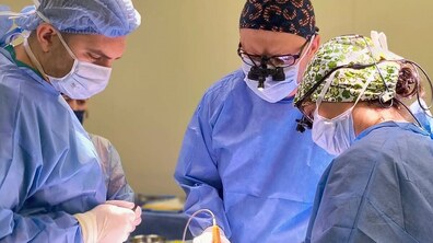 Троє лікарів у медичних халатах, масках та шапочках оперують; один лікар має одягнуті спеціальні операційні окуляри з підсвіткою