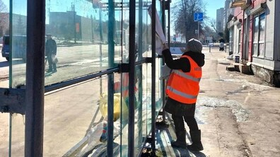 зупинка громадського транспорту зі скляними стінками на міській вулиці. Працівниця комунальної служби у помаранчевому світловідбивному жилеті та сірій в’язаній шапці протирає ганчіркою скло павільйону зупинки. Поруч видно лавку всередині зупинки та пакет із засобами для прибирання. На задньому плані — тротуар, дорога, кілька перехожих і будівлі вздовж вулиці у сонячний зимовий день.