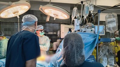 Операційна під час хірургічного втручання: кілька медичних працівників у стерильному одязі, масках і шапочках працюють біля пацієнта під яскравими операційними лампами. Навколо видно медичне обладнання та монітори.