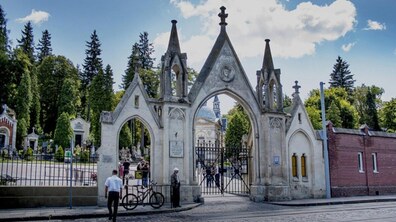 Центральний вхід на територію музею «Личаківський цвинтар» з великою кам'яною брамою з трьома арками. Крізь металеві ворота видно людей, старовинні каплиці, пам'ятники та багато дерев.