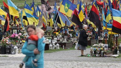 Поле почесних поховань Личаківського кладовища: могили у синьо-жовтих, червоно-чорних прапорах та прапорах різних бригад. Біля однієї із могил стоїть старша жінка, а на передньому плані - молода жінка йде з малям на руках