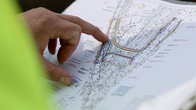 Крупним планом зображено аркуш із технічним кресленням — це план будівництва шляхопроводу. На папері видно складну схему з лініями різних кольорів, підписами, позначками та вигинами дороги чи під’їздів.