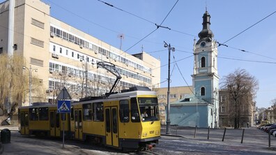 Міський пейзаж із жовтим трамваєм, який рухається вулицею. Над дорогою простягаються трамвайні дроти, а поруч видно знак пішохідного переходу та кілька ліхтарів. На тлі — світла багатоповерхова будівля та церква з високою дзвіницею і годинником.