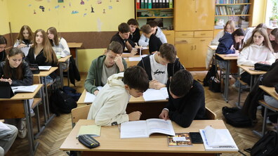 Учні старших класів сидять за партами та працюють із підручниками й зошитами. На передньому плані двоє хлопців схилилися над відкритою книжкою, обговорюючи навчальне завдання. Жовта стіна кабінету прикрашена дитячими малюнками та географічними силуетами, а на задньому плані є шафи з книжковими полицями та папками.
