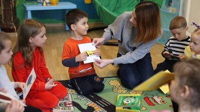 Діти сидять колом на килимі, тримаючи в руках картки із зображеннями. Вихователька спілкується з хлопчиком у помаранчевій футболці, показуючи йому картку з малюнком жовтої машини