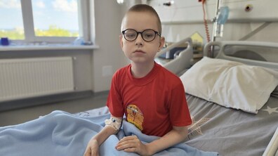 Пацієнт Віталік сидить на лікарняному ліжку у своїй палаті. Він одягнений у червону футболку та має окуляри, на руці – пов’язка після медичної процедури. Поруч видно лікарняне обладнання.