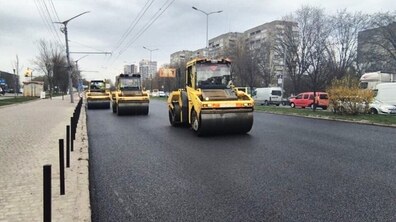 Частина вул. Стрийської під час робіт з асфальтування. Асфальт виглядає новим, рівним і ніби щойно покладеним. По дорозі рухаються три великі дорожні катки — жовті машини для ущільнення асфальту. Вони їдуть один за одним у колоні, займаючи праву частину дороги. Ліворуч — тротуар, відділений від дороги низьким бордюром і рядом чорних металевих стовпчиків. Над дорогою видно дроти для тролейбусів, підвішені між стовпами.