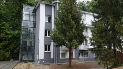 Тимчасове житло на вул. Замарстинівській, яке місто облаштувало для ВПО: триповерхова сіро-біла будівля серед зелених дерев, поруч — пішохідні доріжки та невеликий газон.