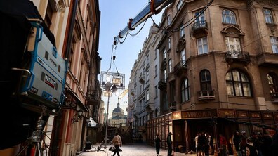 Вузька вулиця з бруківкою та багатоповерховими кам'яницями, де проходять кінозйомки. У центрі кадру стоїть довга стріла підйомного крана, що тримає освітлювальне обладнання над проїжджою частиною. Вздовж вулиці перебувають члени знімальної групи та перехожі.