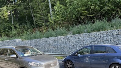 На передньому плані видно два авто – сірий та синій, що рухаються автомобільною дорогою. За металевим відбійником уздовж дороги протягнулася висока підпірна стіна з габіонів — металевих сіток, щільно заповнених сірим колотим каменем. Одразу над кам'яною стіною піднімається схил, густо вкритий зеленим листяним лісом та кущами.