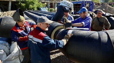 П’ятеро робітників у спецодязі монтують великі чорні ізольовані труби тепломережі на відкритому майданчику. Чоловіки в захисних рукавицях, кепках та зварювальних масках фіксують та зварюють масивні металеві конструкції. На задньому плані видно спецтехніку, насипи ґрунту та житловий будинок.