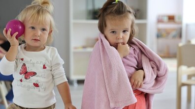 На фото дві дівчинки у садочку. Одна з них тримає м’ячик біля голови, інша - дивиться у камеру, тримаючи палець в роті