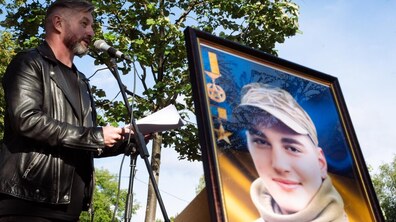 Сергій Жадан виступає з промовою біля мікрофона, тримаючи в руках аркуші з текстом. На передньому плані розміщено велике портретне фото Павла Думанського – наймолодшого львів’янина-Героя України, на тлі синьо-жовтого прапора та з військовими відзнаками.