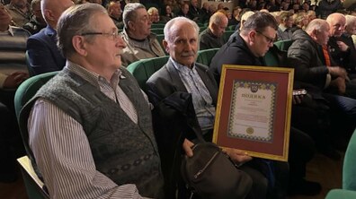 На передньому плані двоє літніх чоловіків сидять на зелених кріслах у залі Львівського палацу мистецтв. Один із них, сивий і з вусами, тримає в руках велику подяку у золотистій рамі з червоним фоном. На документі зверху видно герб України. Позаду них сидять інші люди.