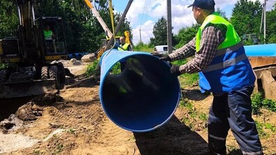Роботи із заміни водогону. На передньому плані працівник у світловідбивному жилеті скеровує велику синю пластикову трубу, яку за допомогою спецтехніки укладають у підготовлену траншею. Позаду працює екскаватор та ще кілька робітників.