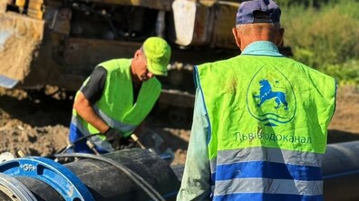 Ремонтні роботи із заміни водопроводу. На передньому плані видно масивні чорні труби та спину працівника Львівводоканалу з логотипом підприємства, а на задньому плані — будівельну спецтехніку та купу землі. Другий робітник у салатовій кепці нахилився над трубами під час виконання роботи.