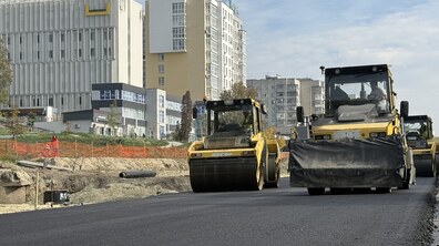 Асфальтування вул. Мазепи. На свіжому чорному асфальті працюють жовті дорожні катки для ущільнення покриття. Ліворуч від дороги видно незавершену земляну ділянку з будівельними матеріалами та металевою огорожею. На задньому плані розташовані високі міські будівлі різних кольорів.