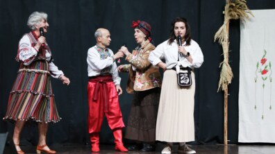 Театральна вистава «Кайдашева сім’я». На сцені виступають четверо акторів старшого віку в яскравих українських народних костюмах. Вони тримають мікрофони та відтворюють діалог між персонажами твору. На задньому плані є чорна сценічна завіса, а праворуч розміщена декоративна композиція зі снопом збіжжя та вишитим рушником.