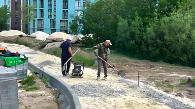 Двоє робітників у парку, серед зелених дерев, облаштовують пішохідні доріжки. Позаду них видніється багатоповерховий новозбудований будинок
