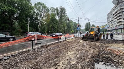 Ремонт тротуару: старе покриття демонтовано й підготовлена основа для нової пішохідної зони. Поруч стоїть жовтий трактор та працівники. Робоча зона відокремлена від проїжджої частини помаранчевою сіткою. Також видно зелені дерева та багатоповерхівку
