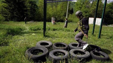 Школяр у військовій формі і захисному спорядженні тренується бігу з перешкодами. Обережно переступає з одного колеса на інше. Довкола зелена місцевість
