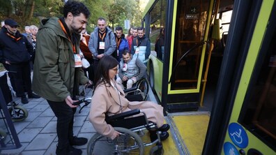 На фото жінка на кріслі колісному заходить в автобус. Чоловік позаду їй допомагає