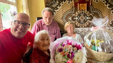 Ювілярка, її родичі та працівник управління соціального захисту усміхаються, позуючи для спільного селфі. Поруч на столі стоїть великий Кошик довголіття з фруктами, а в руках ювілярка тримає красивий букет різнокольорових квітів.