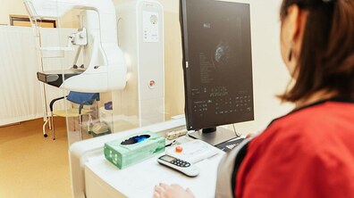 Світлий медичний кабінет із діагностичним обладнанням. Медична працівниця за комп’ютером переглядає результати обстеження. Поруч розташований апарат для мамографії – мамограф.