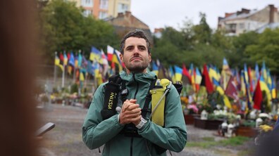 Ультрамарафонець і благодійник Фредерік Сплендор зі спортивним спорядженням стоїть на території Личаківського військового цвинтаря. Він склав руки перед собою та зосереджено і трохи задумливо дивиться вдалечінь. На задньому плані видно багато українських прапорів, які майорять над могилами полеглих військових.