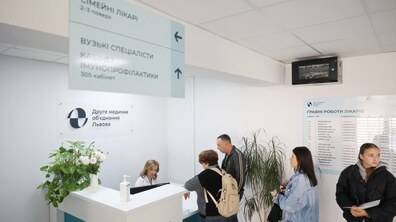 Рецепція медичного закладу. За стійкою адміністратора сидить працівниця у білому халаті та спілкується з відвідувачами. Перед нею стоять троє людей: жінка з рюкзаком нахилилася до стійки, поруч чоловік і ще одна жінка чекають своєї черги. Праворуч стоїть дівчина з документами в руках. На стіні розміщена вивіска з логотипом та написом Друге медичне об'єднання Львова. Також видно інформаційний стенд із графіком роботи лікарів.