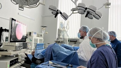 Лікарі у спеціальному медичному одязі оперують пацієнта з допомогою сучасного медичного апарату – гольмієвого лазера, бачачи процес операції на великому екрані; поруч асистує медична сестра