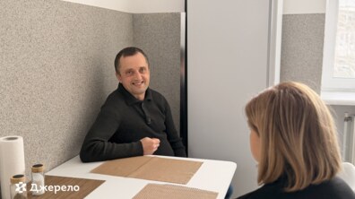 Григорій сидить за столом та усміхається, навпроти нього сидить співрозмовниця; чоловік одягнений у светр темного кольору