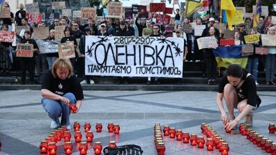 Мирна акція пам'яті, присвячена трагедії в Оленівці, де на передньому плані дві жінки присіли й запалюють червоні лампадки, викладені на бруківці. На задньому плані стоїть група людей із прапорами України та плакатами, головний з яких містить напис «28.07–29.07 Оленівка. Пам'ятаємо, помстимось». Фото з архіву.