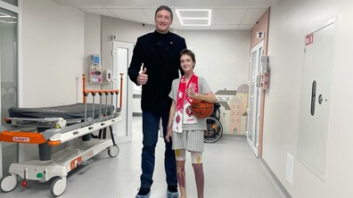 У світлому коридорі медичного закладу стоять двоє людей і позують для фото. Ліворуч — високий чоловік, відомий український баскетболіст Сергій Ліщук. Він одягнений у темне пальто, джинси та бахіли, усміхається і показує жест «палець вгору». Праворуч від нього — дівчинка Настя Цяпало. Вона нижча на зріст, усміхнена, стоїть поруч, притулившись до нього. На Насті світла смугаста лікарняна піжама, на ногах — капці. На її ногах видно післяопераційні пов’язки або медичні бинти. На шиї в неї червоно-білий шарф, а в руках вона тримає баскетбольний м’яч.