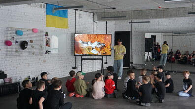 Діти сидять у залі та уважно слухають чоловіка, який проводить для них заняття. Він стоїть біля великого екрана й щось пояснює, а поруч видно український прапор.