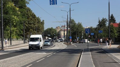 Широка міська вулиця з численними дорожніми знаками. Дорогою їдуть автомобілі, а по тротуарах йдуть пішоходи. Вздовж вулиці видно дерева та ліхтарі.