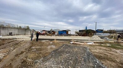 Відкрита територія будмайданчика з нерівним ґрунтом на передньому плані та слідами від важкої техніки. У центрі — прямокутна ділянка. На майданчику працює кілька людей. Позаду металеві контейнери та машини.