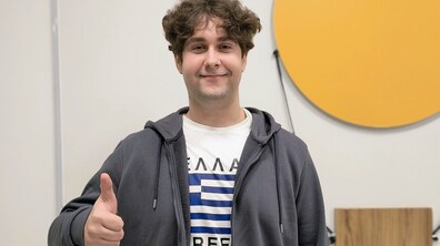 Пацієнт Максим стоїть у холі лікарні й усміхається та показує жест «лайк» великим пальцем. Хлопець одягнений в білу футболку з синьо-чорним принтом і темну кофту з капюшоном. На фоні – світла стіна та круглий жовтий елемент декору.