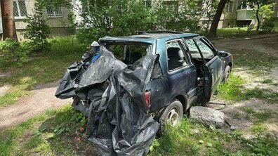 Занедбаний легковий автомобіль із вибитими вікнами, частково накритий чорною плівкою, яка звисає з кузова. Автівка стоїть на прибудинковій території.