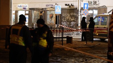 Поліцейські на місці теракту. Позаду огороджувальна стрічка, за якою стоять правоохоронці біля автомобіля рятувальної служби. Внаслідок вибуху на дорозі видно уламки. В магазинах розбиті вітрини та понівечені вивіски.