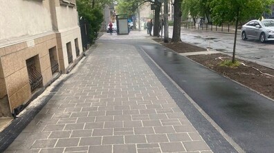 Тротуар зі світло-сірої плитки, що межує з асфальтованою темно-сірою велодоріжкою. Вони розділені тонкою смугою чорної бруківки та пролягають вздовж житлового будинку й дороги. На другому плані видно автівку, ряд дерев, зупинку громадського транспорту та людей