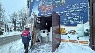 Промислова будівля з великими металевими дверима, які відчинені навстіж. Усередині приміщення видно складені побутові прилади: пральні машини, мікрохвильову піч, холодильник та іншу стару техніку. Ліворуч до входу підходить працівник у темному одязі та червоно-синій куртці з написом комунального підприємства на спині - ЛСКТ. Над входом і праворуч від нього розміщені великі інформаційні банери.