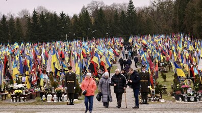 Поле почесних поховань новітніх Героїв Личаківського кладовища у Львові: усі могили - у синьо-жовтих прапорах та прапорах військових бригад, на могилах - квіти та лампадки, над могилами летять птахи; поміж могил ідуть люди, на передньому плані - жінка, двоє літніх людей, чоловік у цивільному та два курсанти військової академії; позаду поля поховань ростуть високі дерева